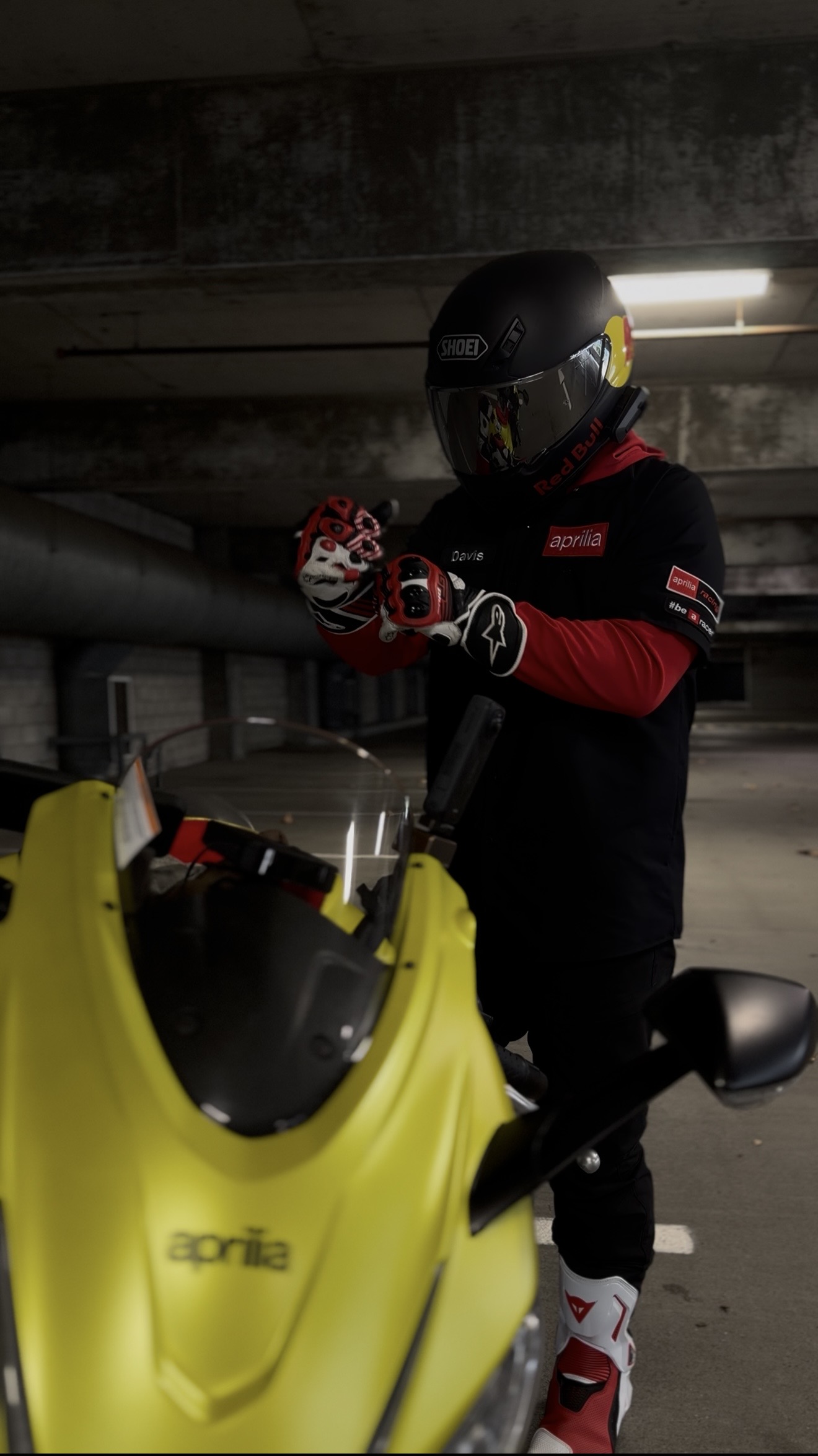 Rider in Aprilia racing gear adjusting gloves next to yellow RS660 in underground parking garage