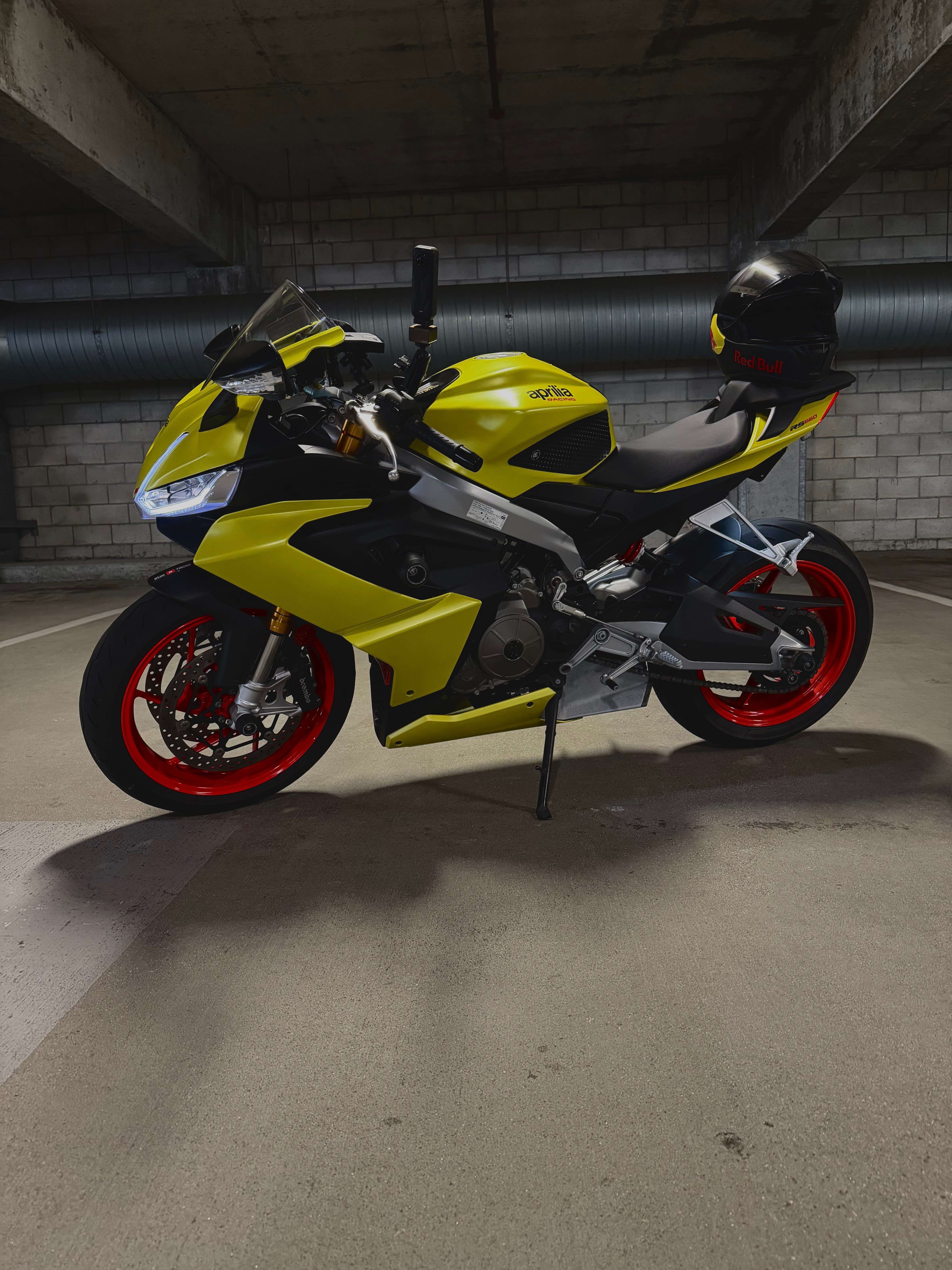 Yellow Aprilia RS660 with red wheels and Red Bull helmet in underground parking garage